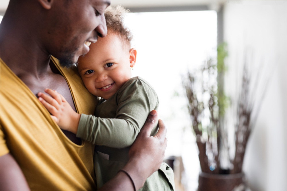 A father holds his young daughter against his chest. She smiles at the camera shyly.