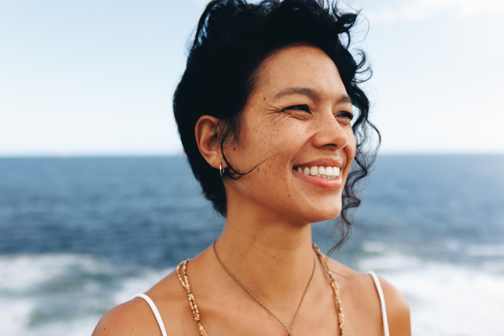 Portrait of a happy woman with wavy hair and bohemian jewelry, smiling at the seaside. Close-up captures her joyful expression.