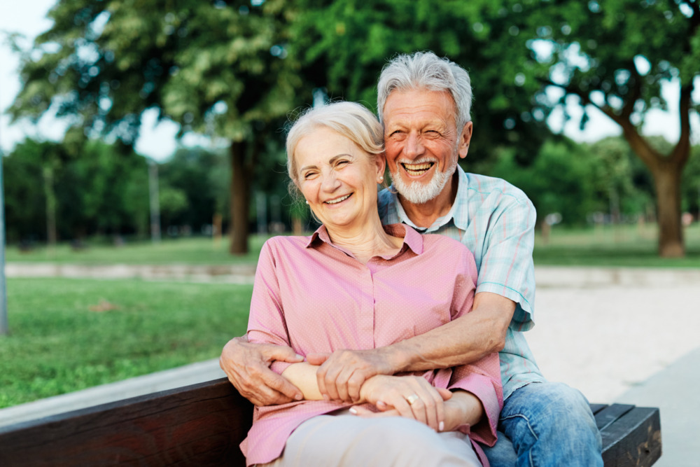 Senior couple sat on a park bench, embracing and smiling.