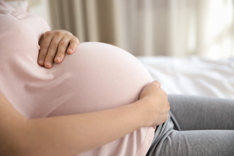 Close‑up image of a pregnant person seated indoors with both hands resting on their abdomen, wearing casual clothing in a softly lit room.