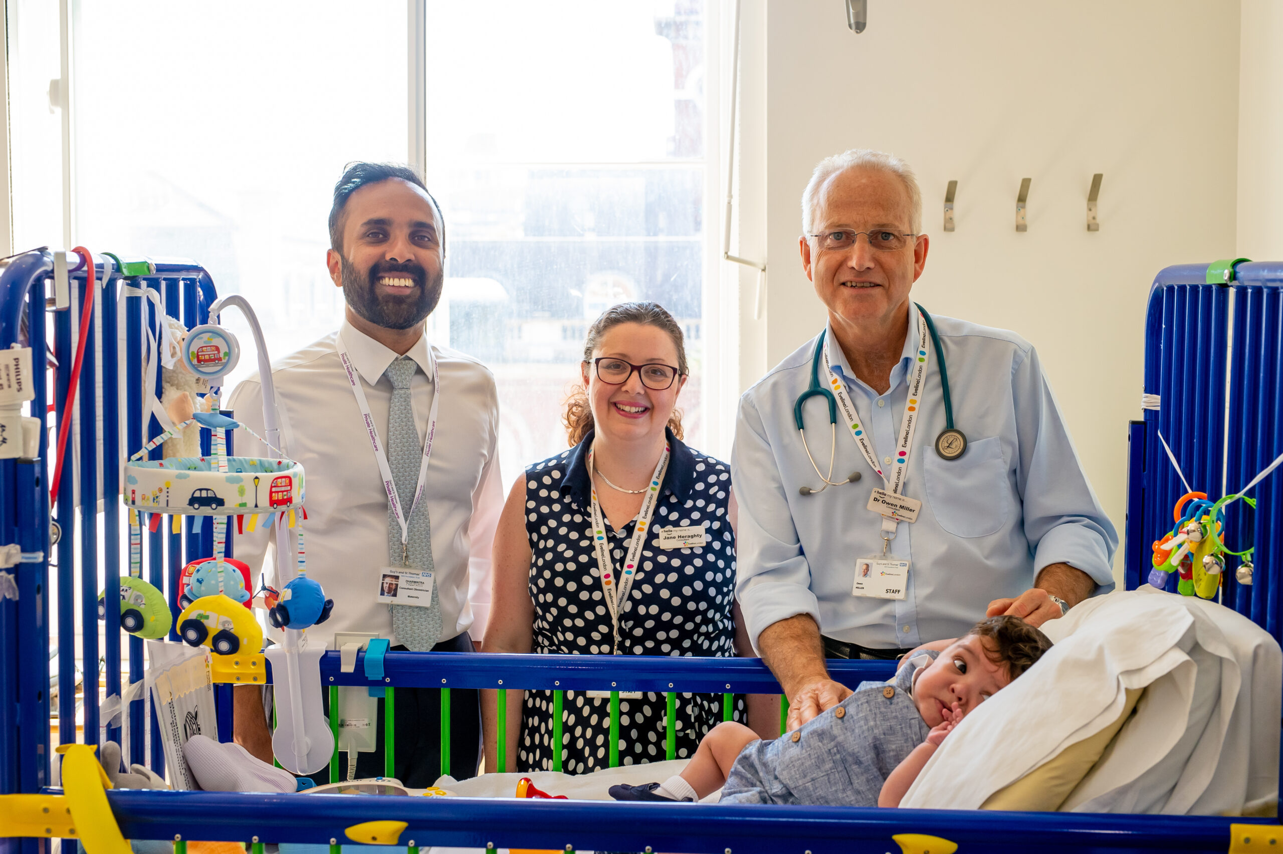 Hospital cot in a clinical ward setting with an infant resting inside, surrounded by medical staff wearing identification lanyards and clinical equipment positioned nearby.