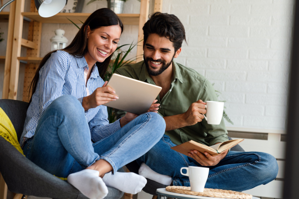 Young couple smiling and looking at a tablet together, drinking coffee. The man is also holding a book.