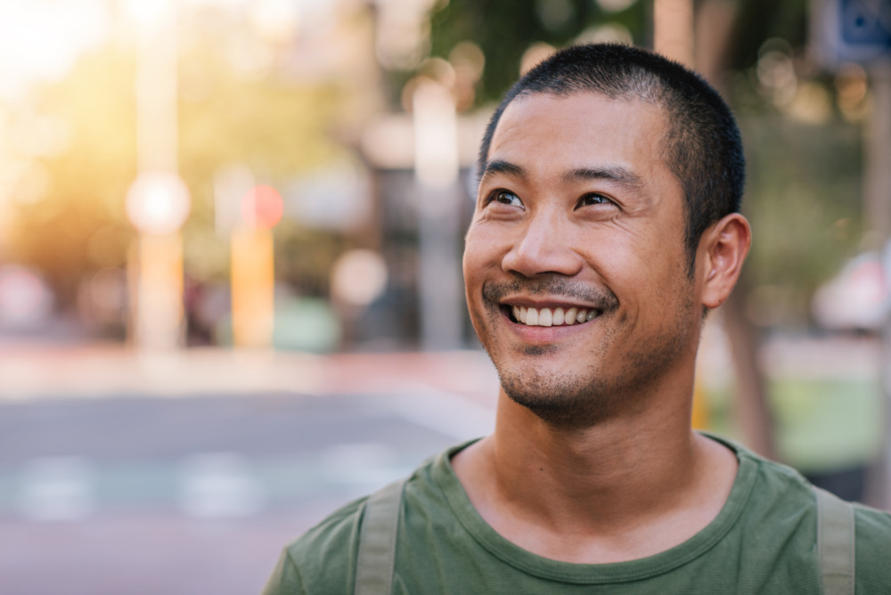 Casually dressed young man looking up and smiling while standing alone outside on a city street on a sunny day