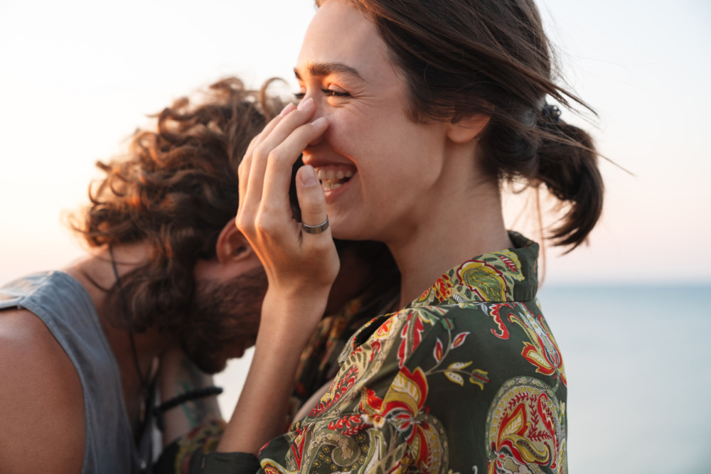 Closeup photo of a couple hugging and laughing on the beach. The woman covers her mouth with her hand and the man rests his forehead on her shoulder.