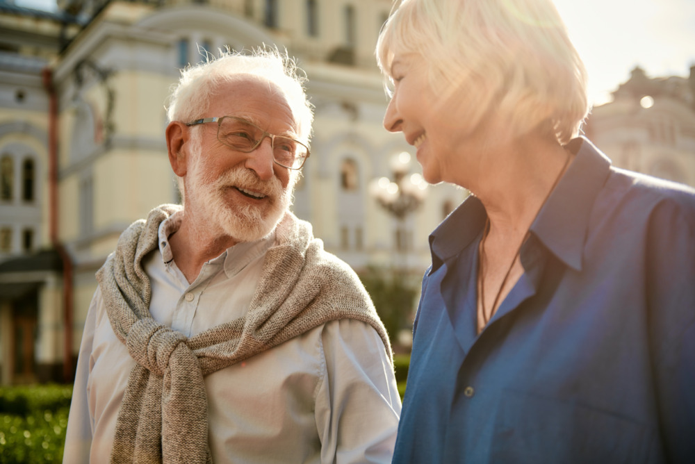 An elderly couple are walking through a city in the sunlight, engaging in happy conversation.