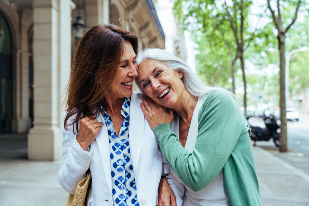 Two mature women walking down the street, laughing together and leaning on each other.