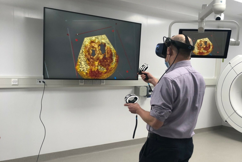 Clinician using a virtual reality headset and handheld controllers to interact with a three‑dimensional medical image displayed on a wall‑mounted screen in a clinical room.