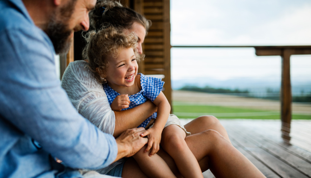 Family with a young daughter sat on a porch. The focus is on the daughter, giggling on her mother's lap.