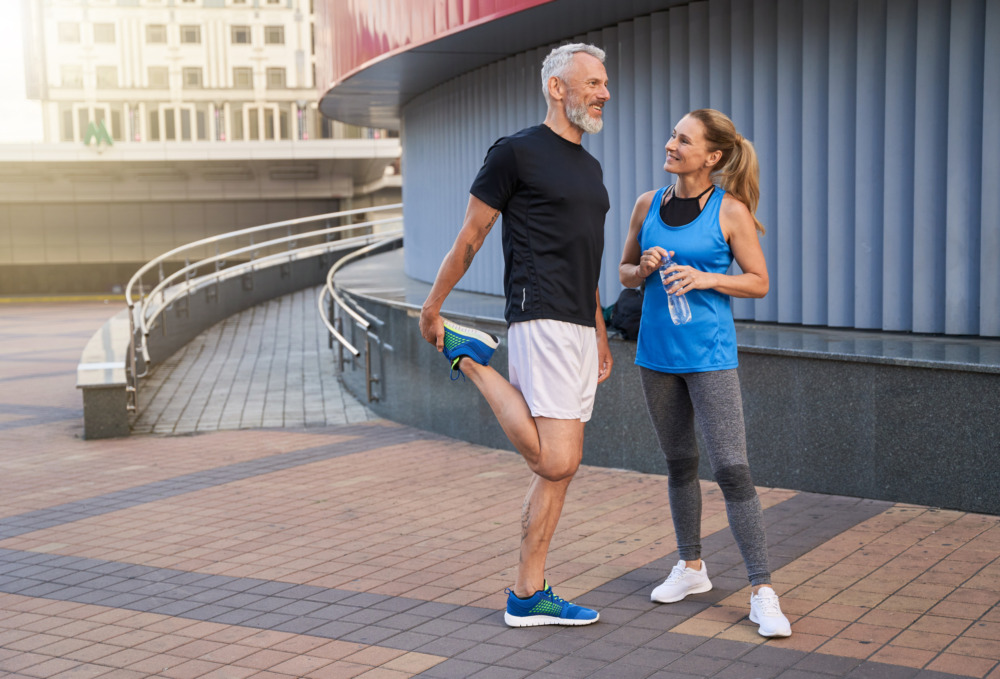A man and woman stand outside a building in conversation. Both are in athletic clothing. The woman tightens the lid of her water bottle and the man stretches his leg.