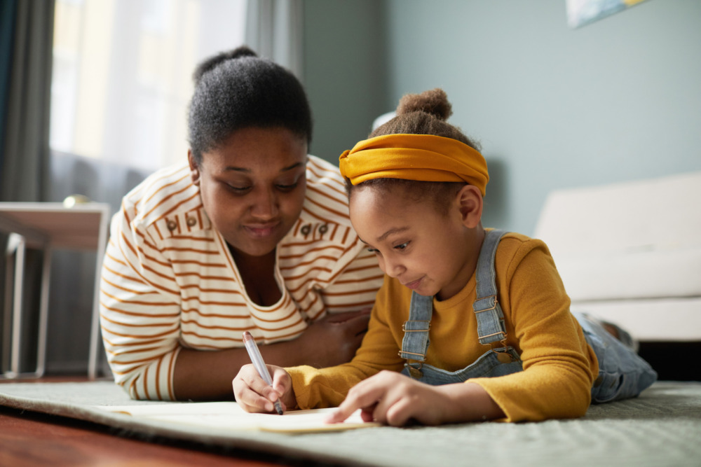 A young girl lying on the floor with her mother, drawing a picture.