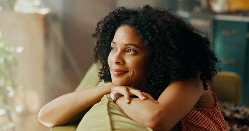 Young woman smiling softly, with her arms folded over the back of a sofa, looking out of a window.