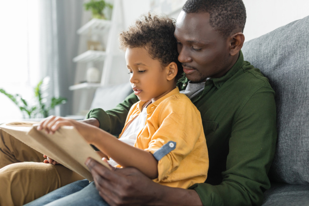 A father and son sit on a sofa reading a book together, with the son sat on the father's lap.