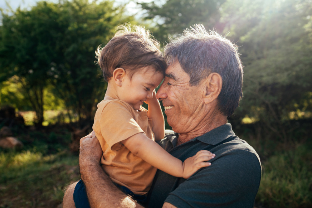 A grandfather holding his grandson outside in the sun. They touch foreheads, and both are smiling.