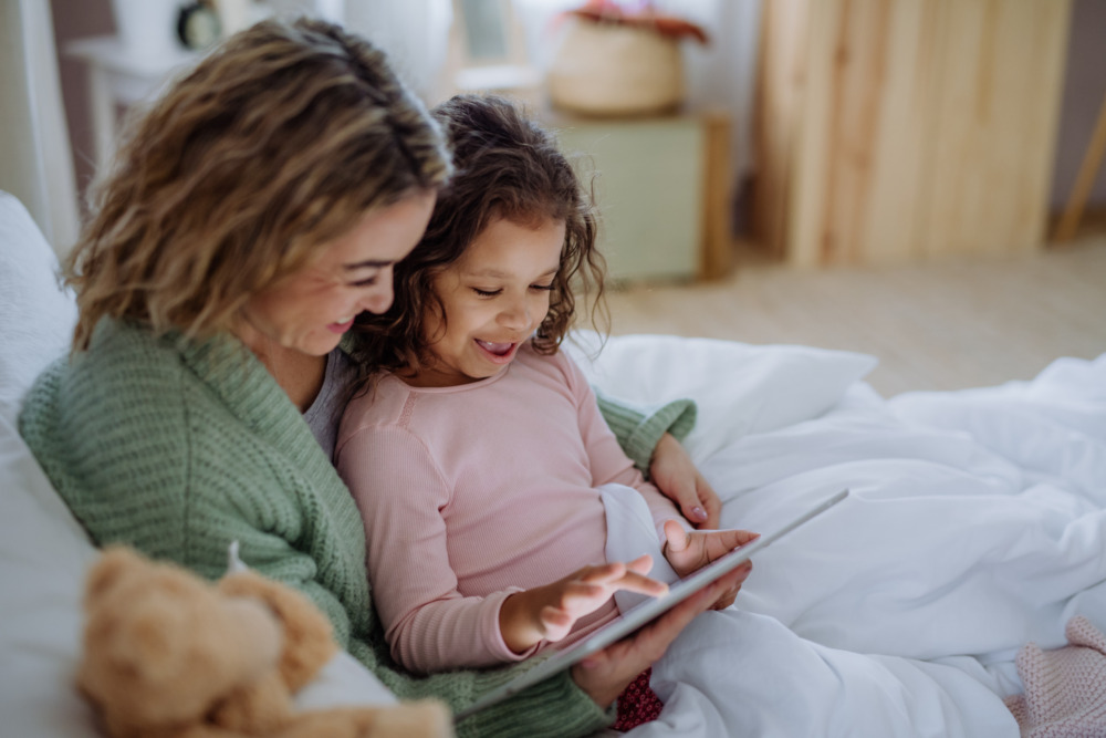 A mother and daughter using a tablet together in bed, and smiling.