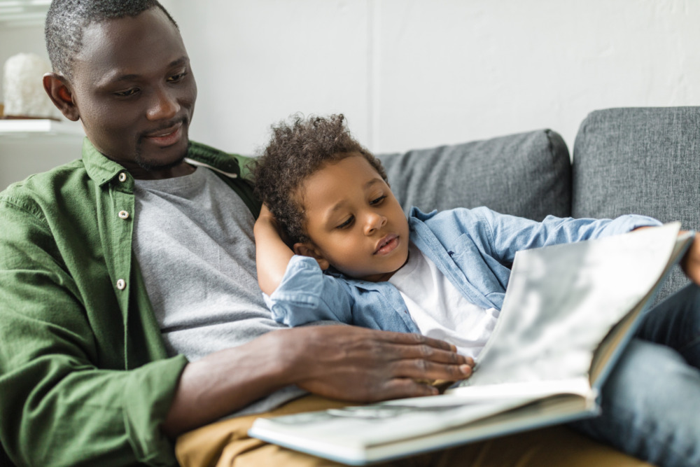 A father reading a storybook with his son, who sits on his lap. They are both lying on the sofa together.