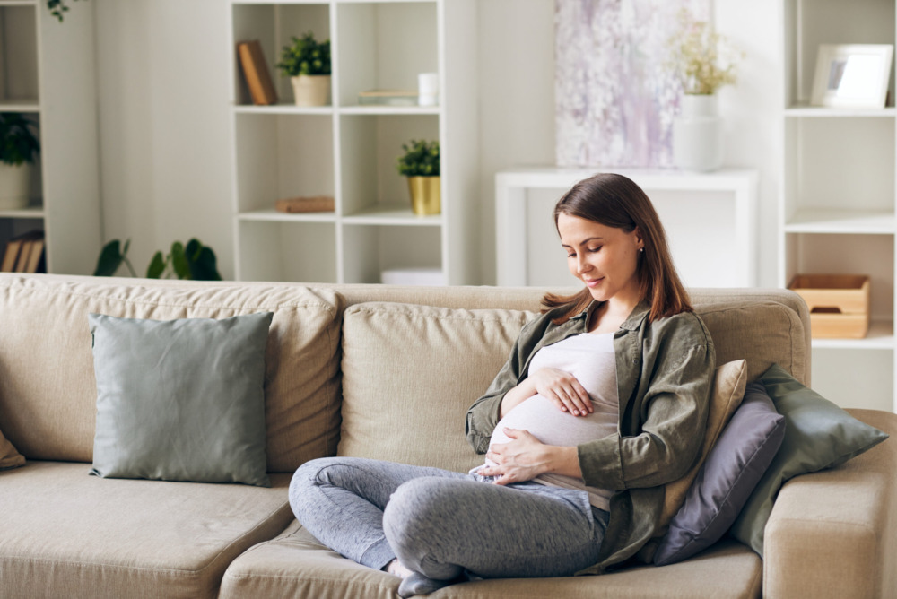 Calm young pregnant woman sitting with crossed legs on sofa and embracing her belly